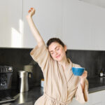 Portrait of happy girl dancing with coffee in the kitchen, wearing bathrobe, enjoying her morning routine